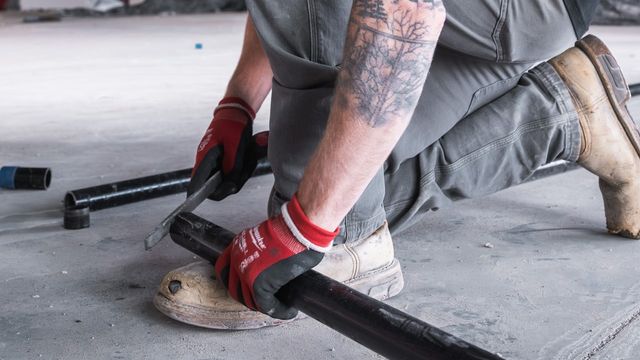 Plumber in work gloves cutting a length of black pipe on a concrete slab